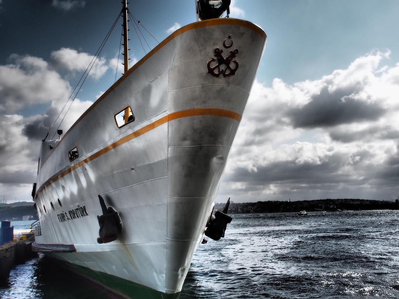 Dramatische Nahaufnahme des Bugs einer klassischen, weiß-grauen Fähre mit gelben Akzentstreifen, die am Steg in Istanbul festgemacht ist. Das Schiff trägt das türkische Emblem (Anker und Halbmond) und den Namen 'FAHRİ S. KORUTÜRK'. Die Szene spielt am Bosporus, einem wesentlichen Bestandteil für jeden 'Bosporus-Flaneure: Mein Guide für einen Spaziergang von Arnavutköy nach Bebek', unter einem stürmischen Himmel mit dramatischen Wolken. Die Uferlandschaft Istanbuls ist dunkel im Hintergrund zu erkennen.