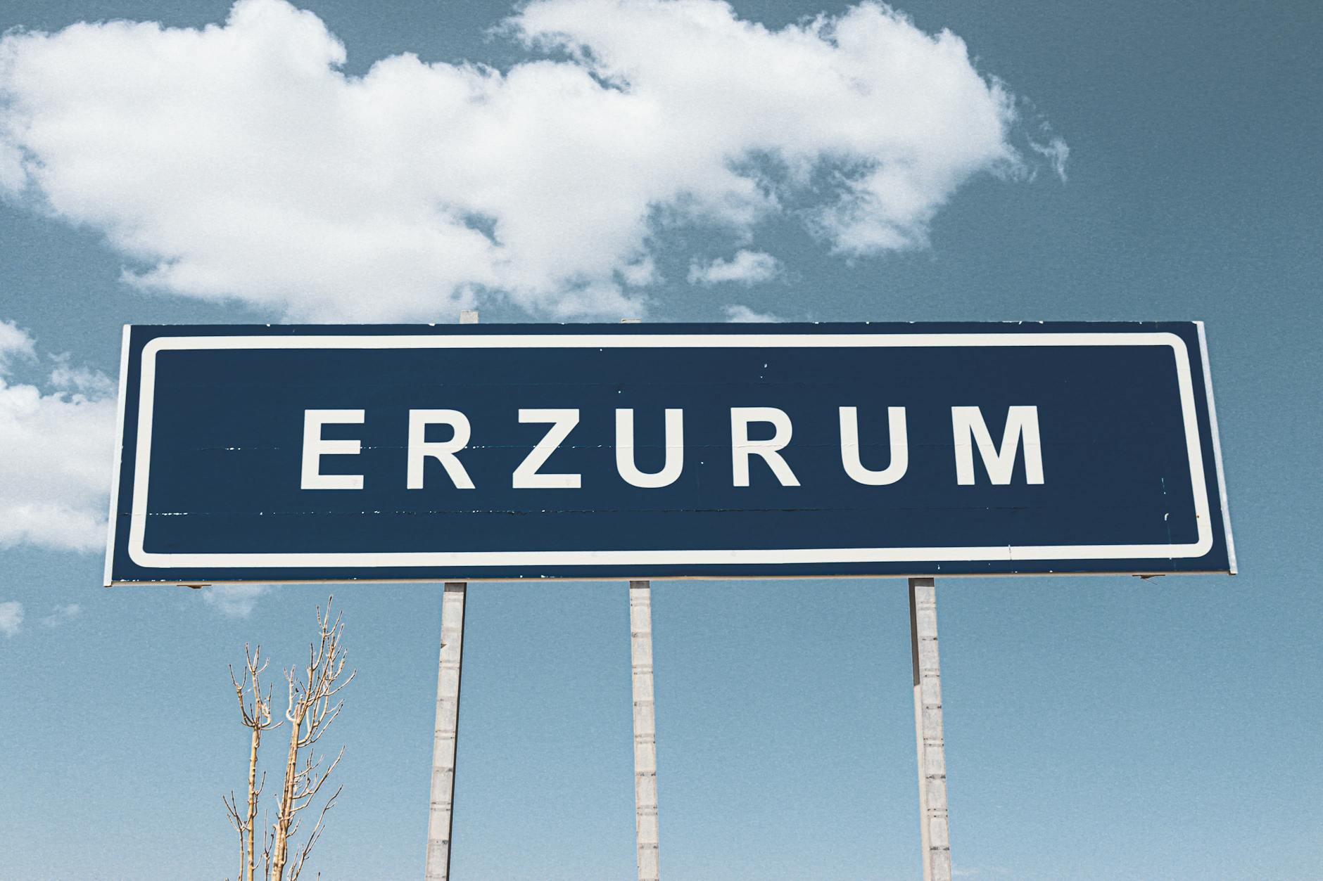 Ein blaues Ortsschild der Stadt Erzurum unter einem leicht bewölkten Himmel in der Türkei.