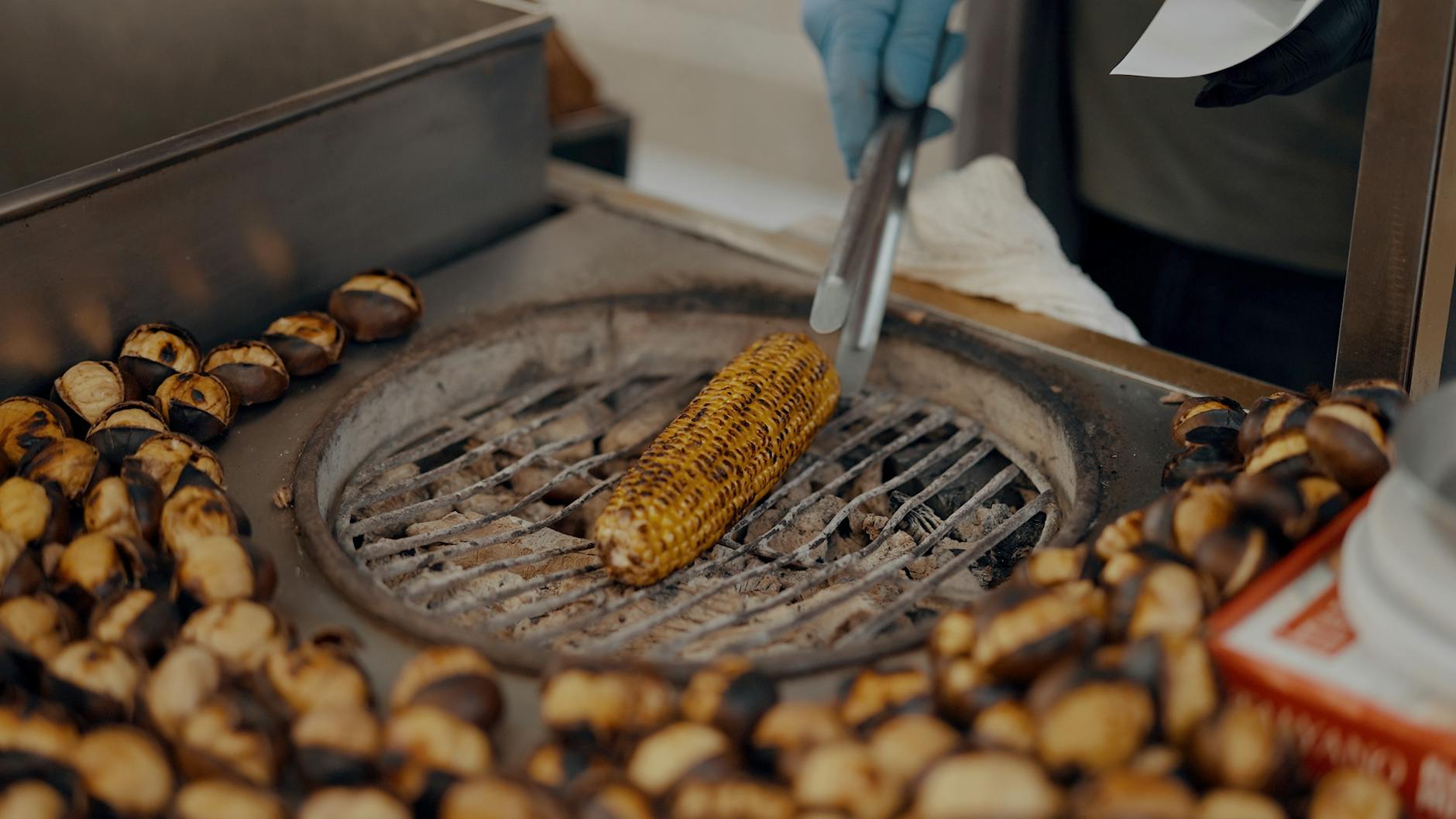 Straßenverkäufer bereitet gegrillten Mais auf Holzkohle zu, umgeben von gerösteten Esskastanien in Istanbul.