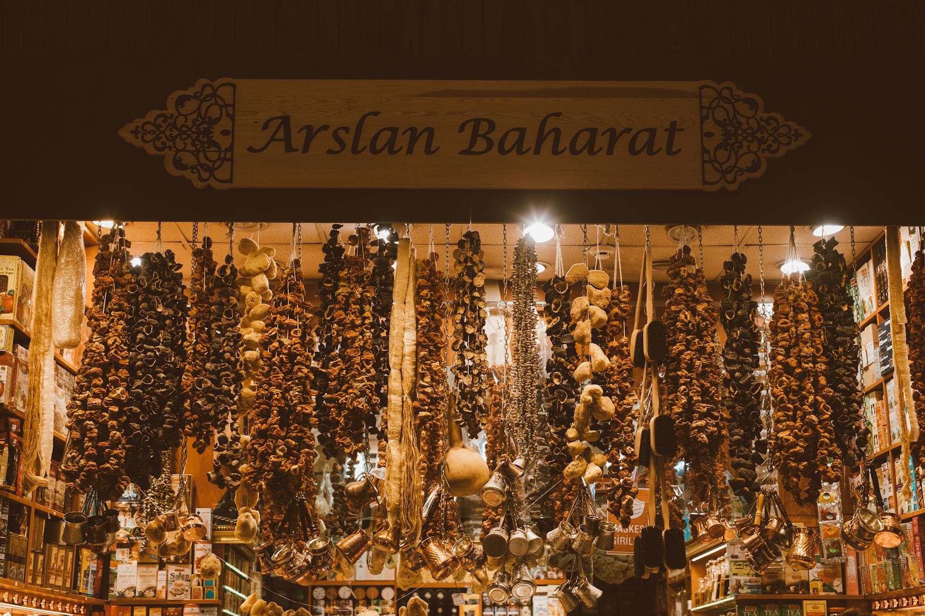 Ein stimmungsvolles Foto aus dem Inneren eines Gewürzladens in Istanbul, möglicherweise dem Großen Basar, das ideal zum Thema "Feilschen wie ein Local: Mein Guide für entspanntes Shoppen auf Istanbuls Basaren" passt. Im Vordergrund hängen reichlich getrocknete Auberginen, Paprika und Schwämme ("kurutulmuş sebzeler") sowie Kupfer-Ibrik-Kännchen herab. Über dem Eingang prangt ein hölzernes Schild mit der Aufschrift "Arslan Baharat", was die authentische Atmosphäre eines türkischen Gewürzladens unterstreicht, wo das Feilschen ein wichtiger Teil des Einkaufserlebnisses ist.