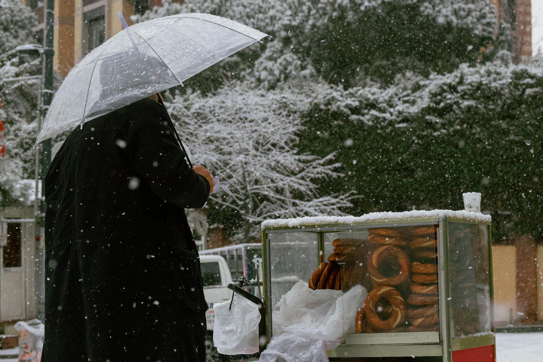 Ein Mann mit Regenschirm kauft Simit von einem Straßenverkäuferwagen bei starkem Schneefall in Istanbul.