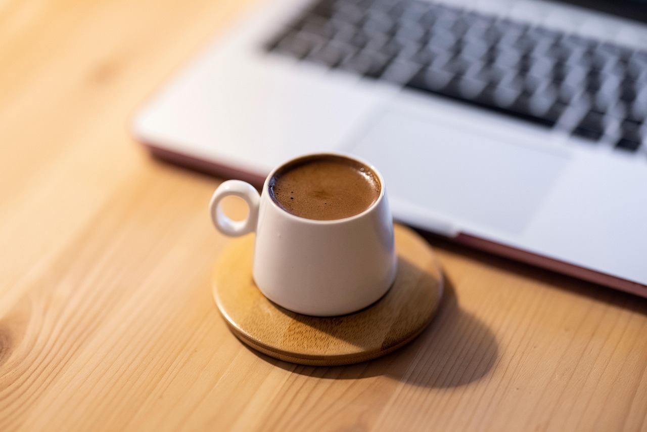 Nahaufnahme einer kleinen, weißen Tasse traditionellen türkischen Kaffees (Kahve) mit dichter Crema auf einem runden Holzuntersetzer, platziert auf einem hellen Holztisch. Im unscharfen Hintergrund ist die Tastatur eines geöffneten Laptops zu erkennen. Dieses Bild symbolisiert die kurze Pause beim Planen oder Schreiben des Artikels "Ehrliche Küche: Mein Guide zu den besten Esnaf Lokantası in Istanbul" und unterstreicht die authentische türkische Komponente durch den Kaffee.