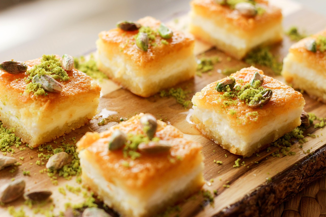 Nahaufnahme von saftigen, quadratischen Stücken eines türkischen Desserts, wahrscheinlich Künefe oder ein ähnlicher süßer Käsekuchen, reichlich beträufelt mit Sirup und bestreut mit gehackten und ganzen Pistazien. Dieses Bild passt gut zu einem Guide über die "Ehrliche Küche: Mein Guide zu den besten Esnaf Lokantası in Istanbul", da traditionelle Süßspeisen ein fester Bestandteil der authentischen türkischen Esskultur sind, die diese Lokantas anbieten.